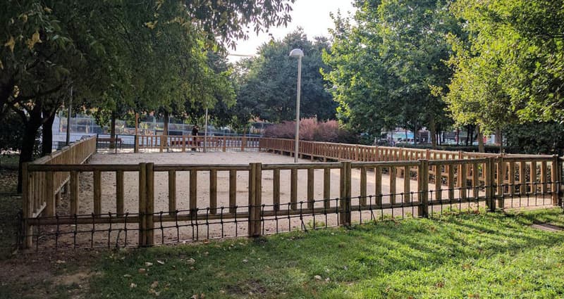 A fenced sand-floored dog run in a Madrid park, bordered by a wooden rail fence and plane trees