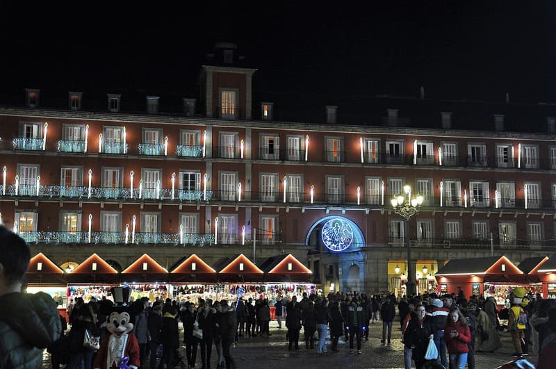 The Plaza Mayor in Madrid on a December night, its arcaded facade lit with candle-shaped lights and a row of red Christmas market stalls below, with a crowd in winter coats crossing the square