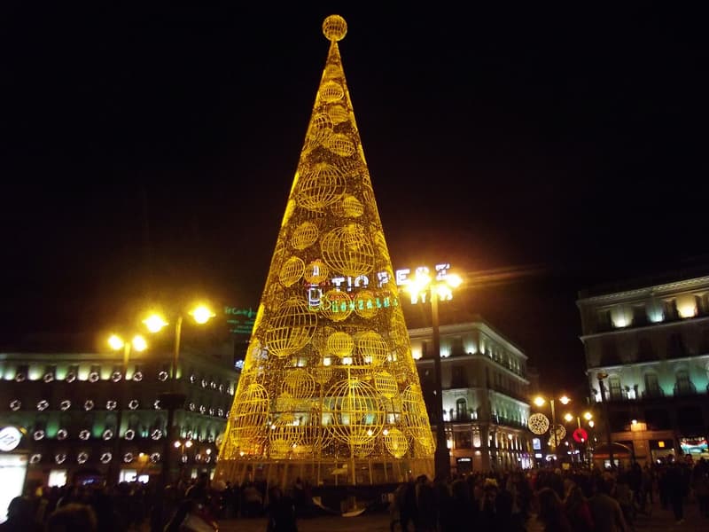 A tall cone-shaped Christmas tree made of gold lights and lit spheres in the Puerta del Sol at night, with illuminated buildings and a crowd around its base