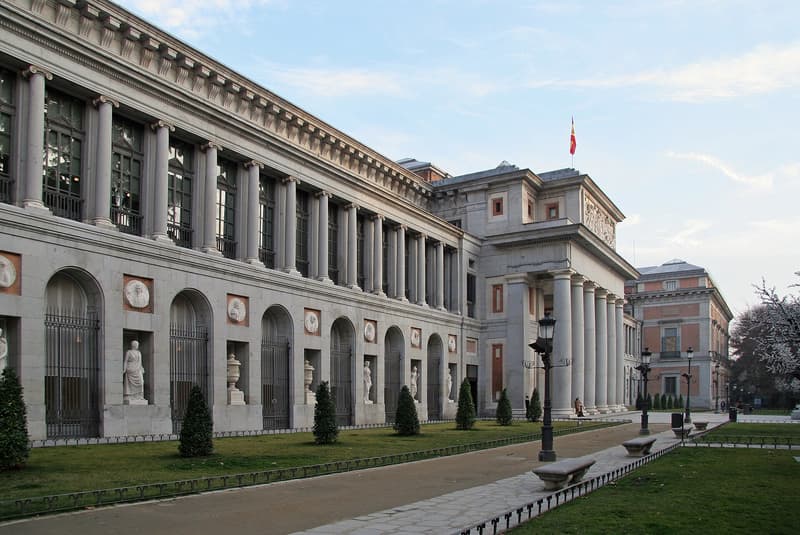 The west front of the Museo del Prado in Madrid, a long neoclassical facade of Ionic columns and arched niches holding statues, with the Velazquez portico and a Spanish flag at the far end