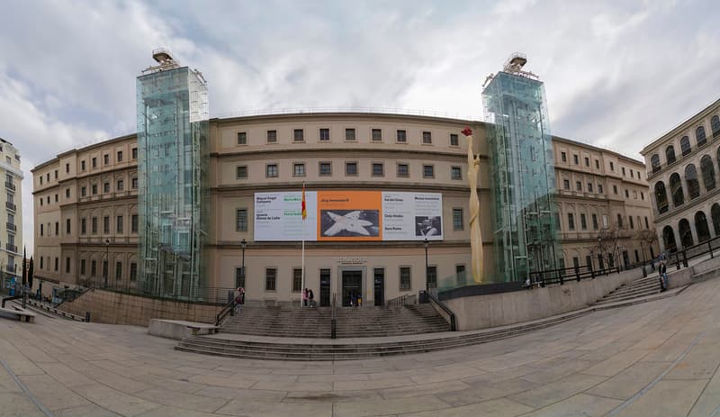 The Museo Reina Sofia in Madrid seen from its plaza, a long stone former hospital building with two external glass and steel lift towers rising at either end and a wide flight of steps in front