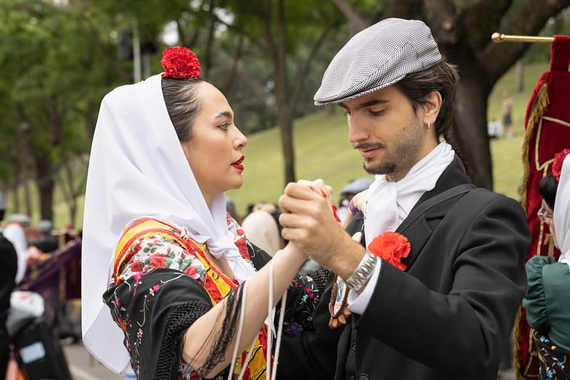 A chulapo in a black suit and flat cap dancing with a chulapa in a white headscarf and red carnations, on a tree-lined path in the Parque de San Isidro during the 2026 fiestas