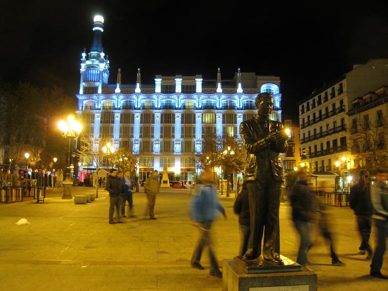 The Plaza de Santa Ana in Madrid at night: a white hotel facade lit in pale blue behind the square, street lamps glowing, a bronze statue in the foreground and people walking past in a blur